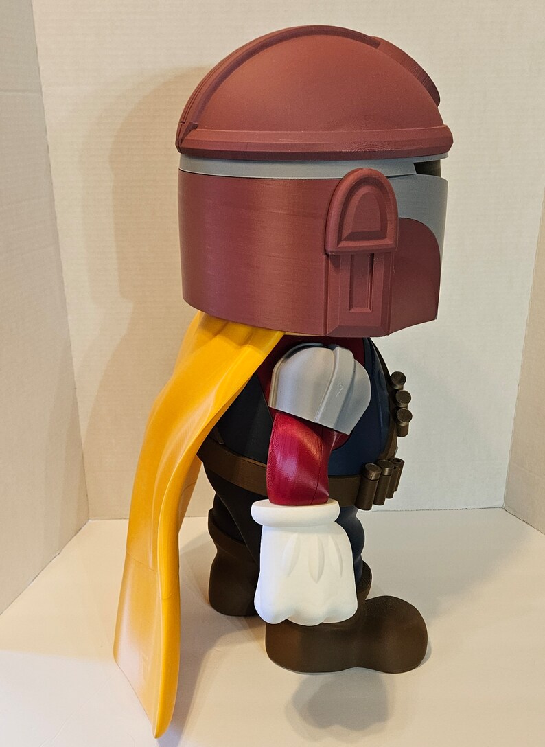 Op de afbeelding: Een rood, wit en grijs 3D-geprint actionfiguur van een personage met een helm, een gele cape en een bruine riem met een bandoleer. Het figuur staat op een wit oppervlak.