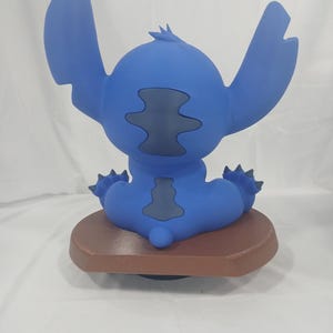 May include: A blue figurine of the character Stitch from the movie Lilo & Stitch, viewed from the back. The figurine has large ears, a small tail, and is sitting on a brown base. The figurine is a solid blue color with darker blue accents.