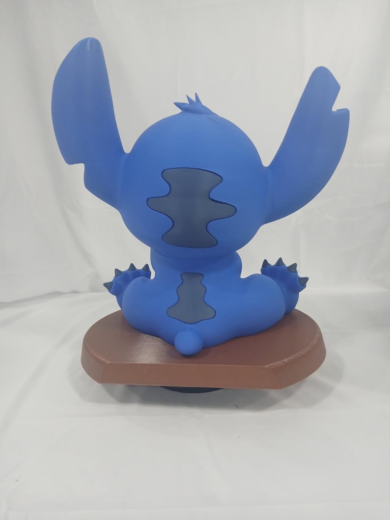 May include: A blue figurine of the character Stitch from the movie Lilo & Stitch, viewed from the back. The figurine has large ears, a small tail, and is sitting on a brown base. The figurine is a solid blue color with darker blue accents.