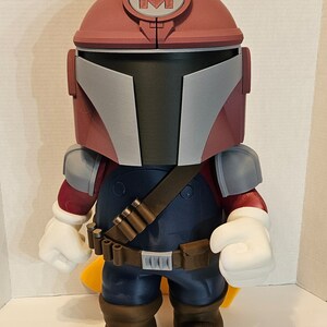 Op de afbeelding: Een rood en grijs speelgoedfiguur met een helm met een grote "M" erop. Het figuur draagt een blauwe jumpsuit met een bruine riem en een bandoleer. Het figuur heeft witte handschoenen en bruine laarzen.