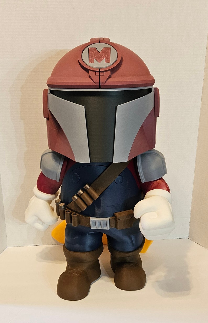 Op de afbeelding: Een rood en grijs speelgoedfiguur met een helm met een grote "M" erop. Het figuur draagt een blauwe jumpsuit met een bruine riem en een bandoleer. Het figuur heeft witte handschoenen en bruine laarzen.