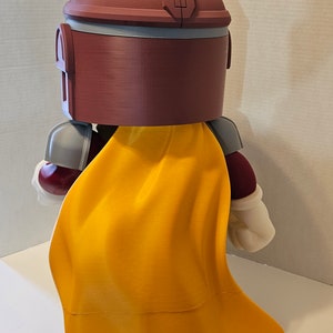 Op de afbeelding: Een 3D-geprint figuur van een personage in bordeauxrood en grijs met een gele cape. Het figuur heeft een helm met een vizier en houdt een wapen vast.