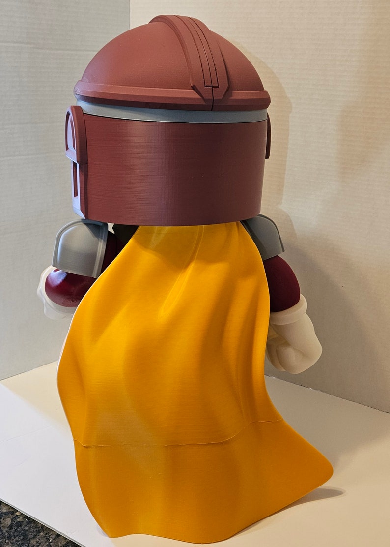 Op de afbeelding: Een 3D-geprint figuur van een personage in bordeauxrood en grijs met een gele cape. Het figuur heeft een helm met een vizier en houdt een wapen vast.