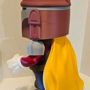 Op de afbeelding: Een 3D-geprint beeldje van een personage met een roodbruine helm, een gele cape en bruine laarzen. Het beeldje houdt een witte handschoen uit.