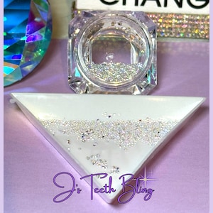 May include: A clear, faceted jar filled with small, sparkling rhinestones sits above a white, triangular tray also containing rhinestones. The tray is on a purple surface. The text "J's Teeth Bling" is written in a cursive font.