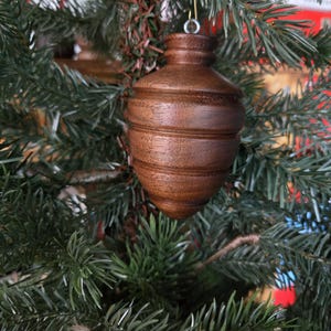 May include: A brown wooden Christmas ornament with a bulbous shape and horizontal grooves. The ornament hangs from a gold-colored wire hook and is set against a backdrop of green Christmas tree branches.