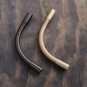May include: Two flexible metal gooseneck pipes, one black and one gold, are shown on a wooden surface. The pipes are bent into a curved shape.