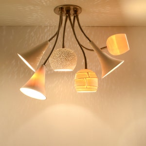 May include: A modern ceiling light fixture with six white and cream lampshades. The lampshades are in various shapes, including round, cone, and rectangular. The light fixture is made of metal and has a brushed nickel finish.