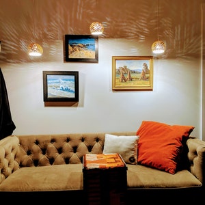 Three 6 inch asymmetrical pendants hang at varying heights above an ochre vintage velvet couch. Patterned projections seem to dance on the wall behind. The scene is cozy and invites the viewer to grab a glass of wine or cup of hot tea and settle in.