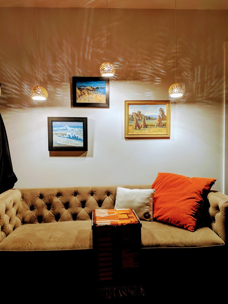 Three 6 inch asymmetrical pendants hang at varying heights above an ochre vintage velvet couch. Patterned projections seem to dance on the wall behind. The scene is cozy and invites the viewer to grab a glass of wine or cup of hot tea and settle in.