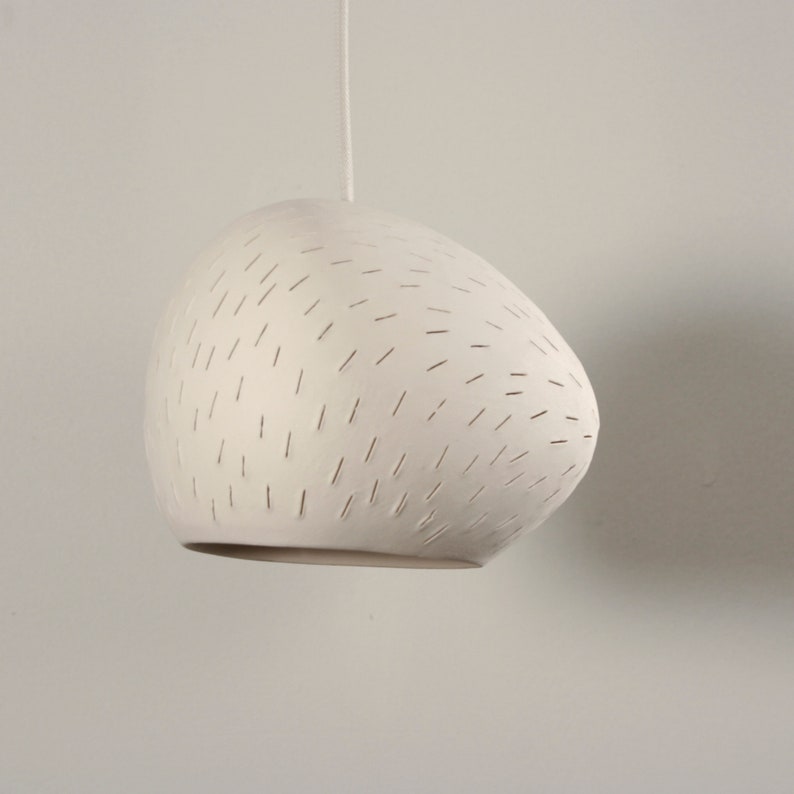 This close-up product shot show cases the lamp when it is off. The matte warm white ceramic's slight imperfections evidence the human hand's role in it's creation. Line perforations sweep across it's surface like a gently current.