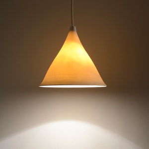 May include: A simple, off-white, cone-shaped lampshade hangs from the ceiling, casting a warm, yellow light onto the wall below.