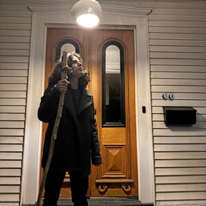 May include: A young person wearing a dark green jacket and black pants stands in front of a wooden door. They are holding a long, brown stick and looking up at a white light fixture hanging from the ceiling.