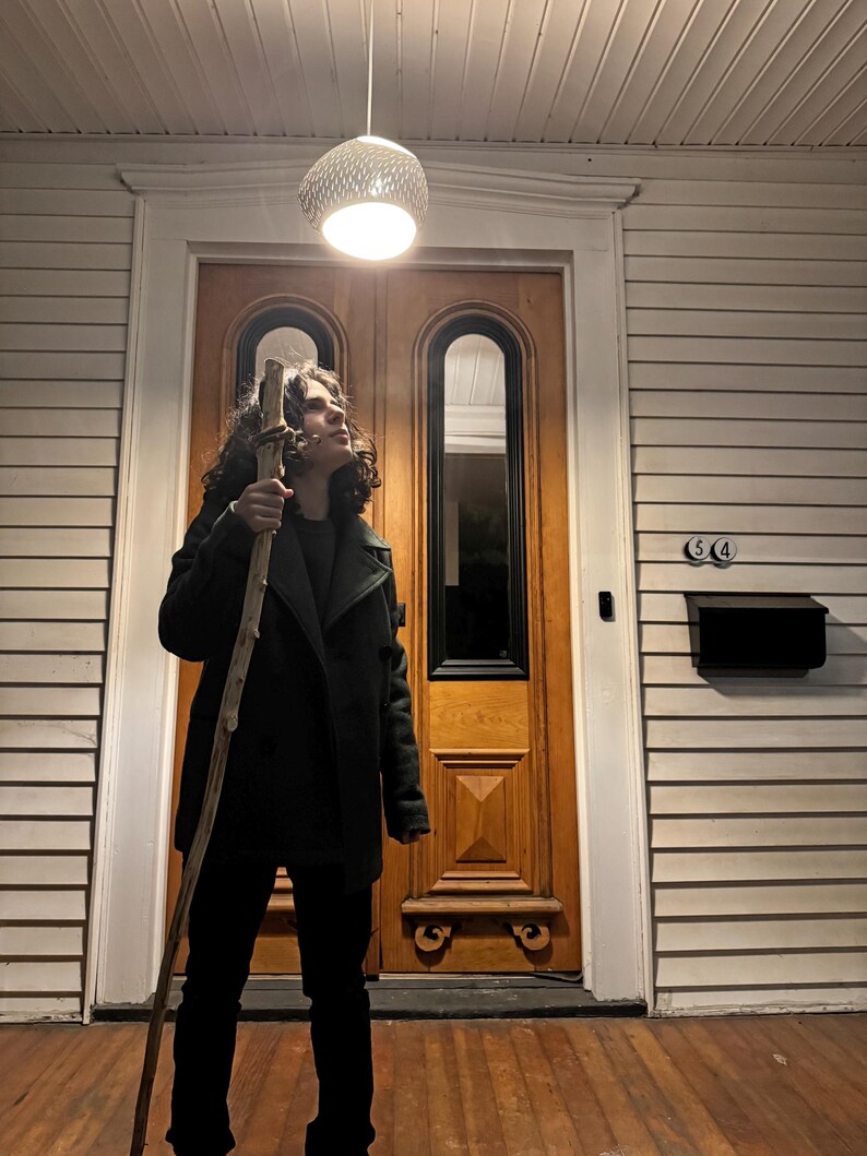 May include: A young person wearing a dark green jacket and black pants stands in front of a wooden door. They are holding a long, brown stick and looking up at a white light fixture hanging from the ceiling.