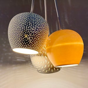 May include: Three hanging light fixtures with a modern design. The fixtures are made of ceramic and have a white, grey, and orange colour scheme. The fixtures have a unique pattern of holes throughout the surface.