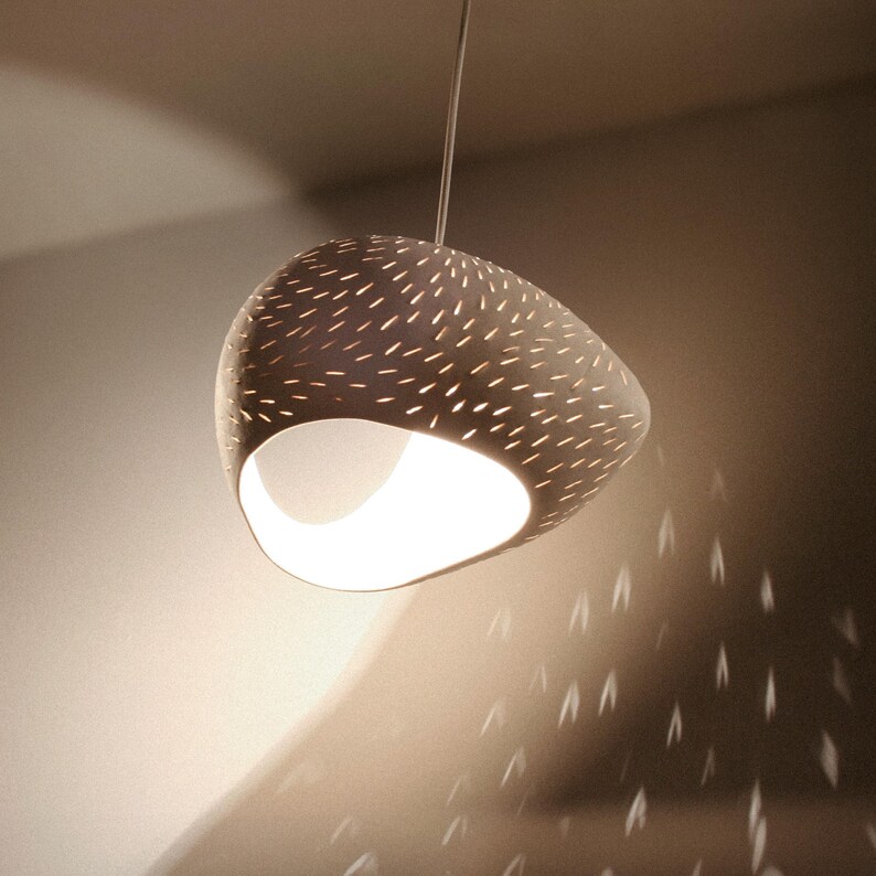 May include: A white ceramic pendant light fixture with a unique, organic shape and a pattern of small holes. The light casts a warm glow and creates interesting shadows on the wall.