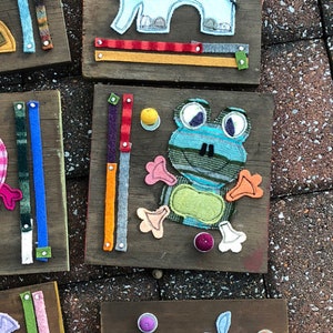 May include: Five wooden boards with felt animal appliques and colorful buttons. Each board has a different animal: a giraffe, an elephant, a frog, a hippopotamus, and a whale. The boards are designed for children to learn about animals and colors.