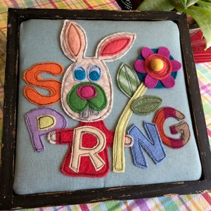 May include: A framed felt wall art piece with the word "SPRING" spelled out in colorful letters. A white bunny with a red shirt and a green nose is holding a yellow flower with a pink center.