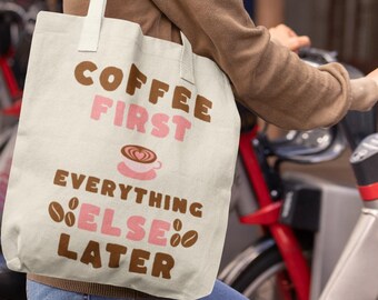 May include: Cream-colored tote bag with the text "COFFEE FIRST EVERYTHING ELSE LATER" in brown and pink. The bag features a coffee cup graphic. The person is wearing a brown top and blue jeans.