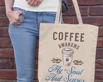 May include: Beige tote bag with the text "COFFEE AWAKENS My Soul And Senses" and a coffee cup illustration. The bag is held by a person wearing blue jeans and a white top, standing in front of a brick wall.
