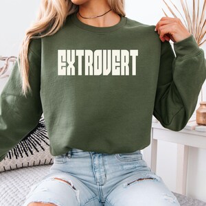 Może przedstawiać: Zielona bluza w kolorze oliwkowym z białym napisem "EXTROVERT" na piersi. Bluza z okrągłym dekoltem jest zestawiona z jasnoniebieskimi, podartymi dżinsami. Obraz prezentuje swobodny, codzienny styl, z dobrze widocznym tekstem.