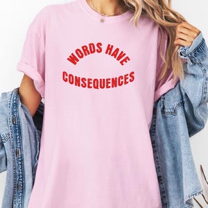 Puede incluir: Camiseta rosa claro con el texto rojo "WORDS HAVE CONSEQUENCES" en un diseño curvo. La camiseta tiene mangas cortas y un corte holgado. Una chaqueta vaquera está sobre los hombros.