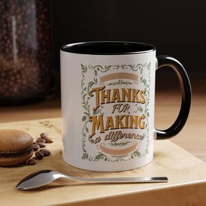 May include: White ceramic mug with a black interior and handle. The mug features the text "THANKS FOR MAKING a difference" in gold and green, with decorative floral elements. A spoon and a macaron are on a wooden surface.