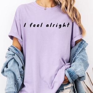 Puede incluir: Una camiseta de color lila claro con la frase "I feel alright" impresa en negro. La camiseta tiene mangas cortas y un corte holgado. La modelo lleva una chaqueta vaquera encima.