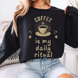 May include: Black crew neck sweatshirt with a gold graphic of a coffee cup and the text "COFFEE is my daily ritual". The design includes coffee beans and star accents. The sweatshirt is worn by a person wearing blue jeans.