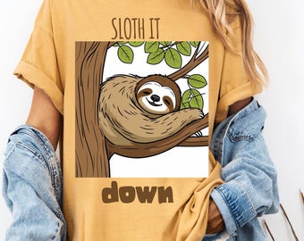 Pode incluir: Camiseta mostarda com um desenho de preguiça de desenho animado num galho de árvore. O texto "Sloth It Down" está impresso na camiseta. A camiseta tem mangas enroladas e um corte descontraído.
