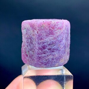 Puede incluir: Una gema de rubí en bruto, sin cortar, de forma cilíndrica, con un tono púrpura rosado intenso. La gema está montada sobre un soporte de exhibición transparente y rectangular, mostrando su textura y color naturales sobre un fondo oscuro. Un espécimen mineral único.