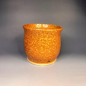 May include: A handcrafted ceramic pot with a textured, speckled surface in shades of orange and brown. The pot has a rounded body and a slightly flared rim. It sits on a light-colored base, showcasing a unique, artisanal design.