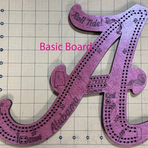 Puede incluir: Un tablero de juego de madera rosa en forma de la letra "A", con las palabras "Roll Tide!" y "Alabama" grabadas. Tiene agujeros para un juego, con las palabras "Start", "Games" y "End". También hay números.