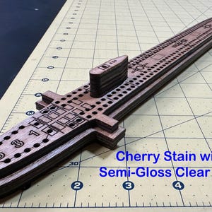 Könnte beinhalten: Ein hölzernes Cribbage-Spielbrett in U-Boot-Form mit Kirschholzbeize und halbglänzender Klarlackierung. Das Brett enthält eingravierte Zahlen und eine Reihe von Löchern für das Spiel sowie einen erhöhten Turmaufbau auf der Oberfläche.