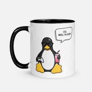 May include: White ceramic mug with a black rim and handle. The mug features a cartoon penguin wearing sunglasses and holding a video game controller and a pink drink. The text "GG, WELL PLAYED" is in a speech bubble above the penguin.