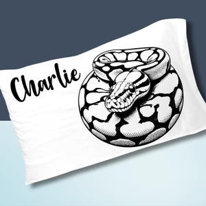 May include: White pillowcase featuring a black and white illustration of a coiled snake. The snake's head is in the center, with the name "Charlie" in cursive script on the upper left.