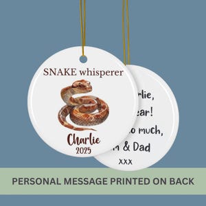 May include: A white ceramic ornament with a watercolor illustration of a snake. The ornament has the text "SNAKE whisperer" and "Charlie 2025". The back of the ornament has a personal message printed on it.