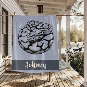 May include: A light blue towel with a black and white illustration of a coiled snake. The towel has a dark gray band at the bottom with the name "Johnny" in a cursive font.