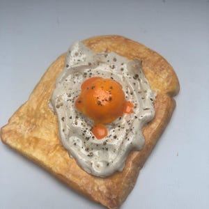 May include: A slice of toast with a fried egg on top. The toast is a golden brown colour, and the egg has a bright orange yolk and a white, speckled egg white. Black pepper is sprinkled on the egg. A close-up image.