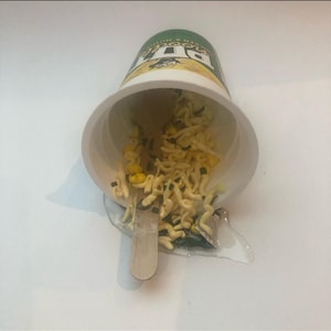 May include: A white cup with a green and yellow label is tipped over, spilling noodles and corn kernels onto a white surface. A wooden stick is lying next to the cup.