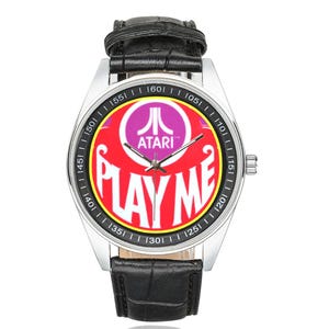 Op de afbeelding: Een zilveren polshorloge met een zwarte leren band. De wijzerplaat heeft een rood en geel ontwerp met het Atari-logo en de woorden "PLAY ME". Het horloge heeft een klassieke analoge weergave.