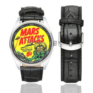 May include: A wristwatch with a black leather band and a silver-colored case. The watch face features a yellow background with the words "MARS ATTACKS" in red, along with a retro alien graphic. The watch band is 9 inches long.