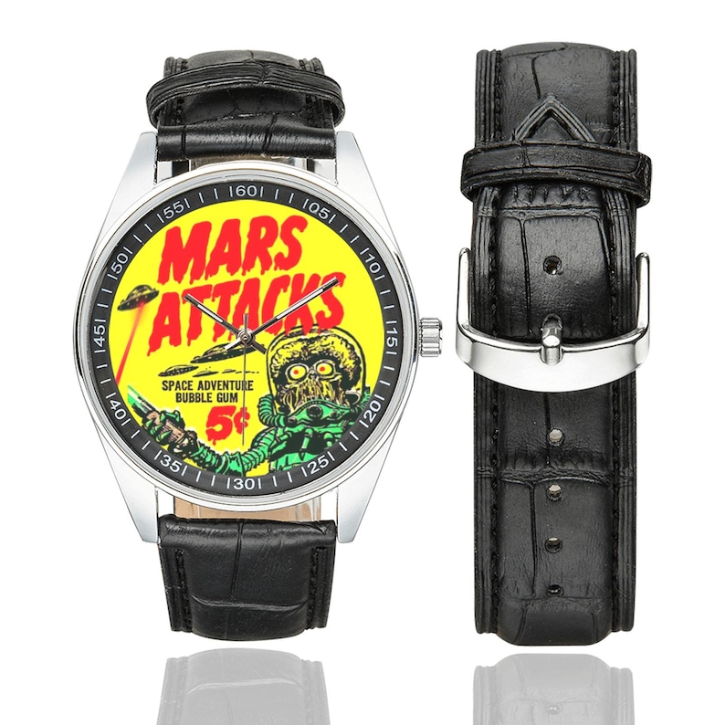 May include: A wristwatch with a black leather band and a silver-colored case. The watch face features a yellow background with the words "MARS ATTACKS" in red, along with a retro alien graphic. The watch band is 9 inches long.