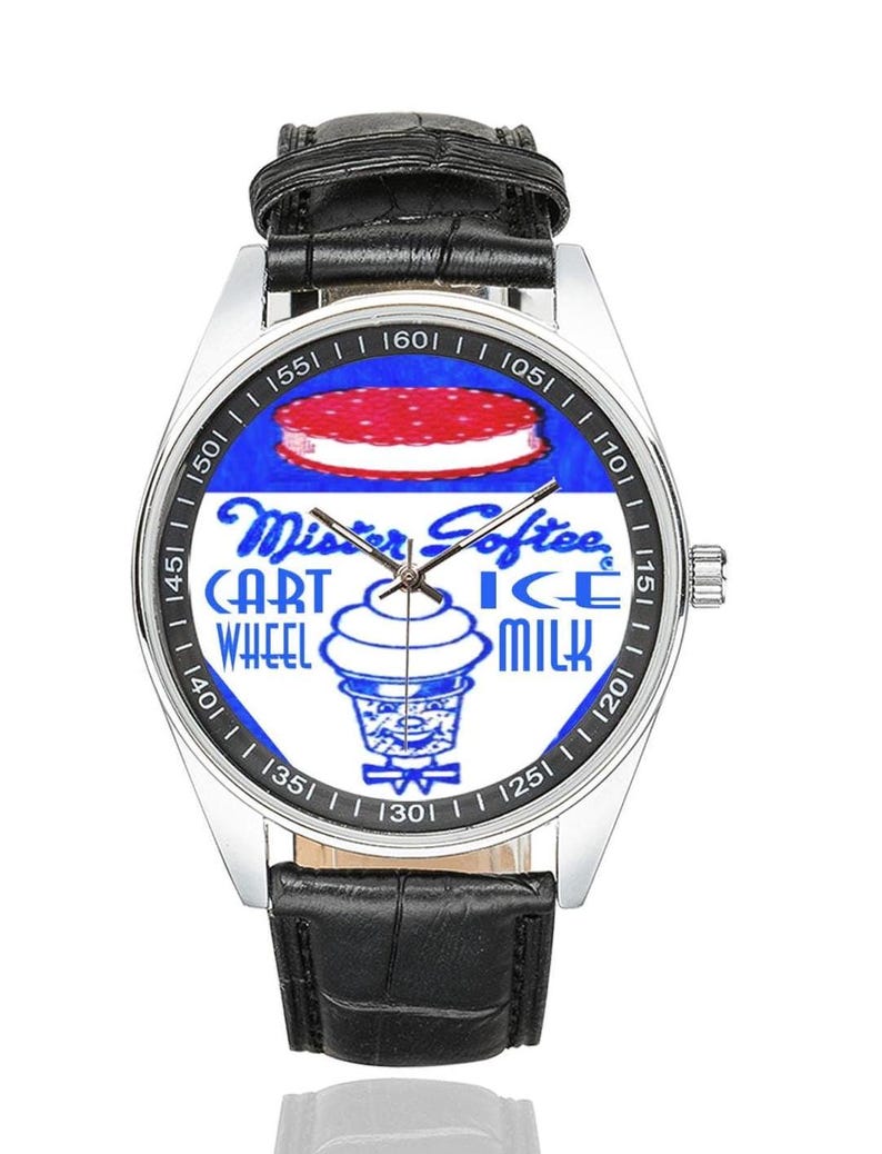 K&ouml;nnte beinhalten: Silberne Armbanduhr mit schwarzem Lederarmband. Das Zifferblatt zeigt ein blau-wei&szlig;es Design mit den Worten "Mister Softee" und "Cart Ice Milk Wheel". Die Uhr hat eine silberne L&uuml;nette und ein schwarzes Lederarmband.