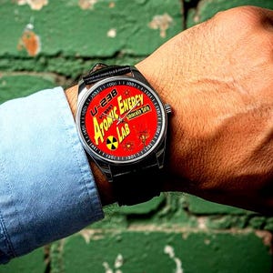 May include: A wristwatch with a black leather strap and a silver case. The watch face is red and features the words "Atomic Energy Lab" and a radiation symbol. The watch is on a person's wrist.