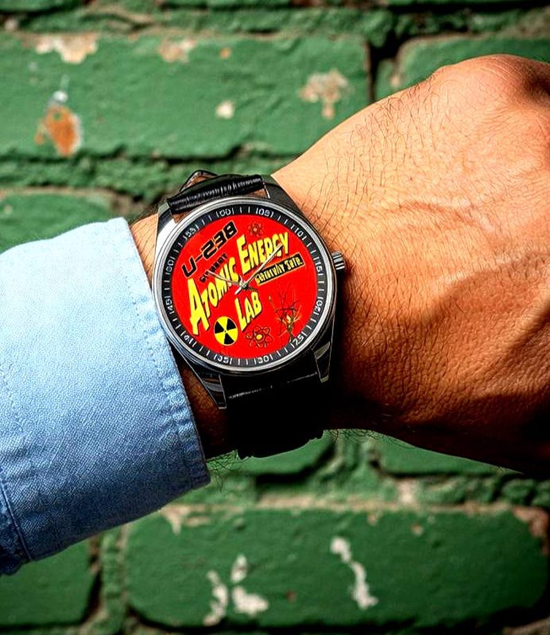 May include: A wristwatch with a black leather strap and a silver case. The watch face is red and features the words "Atomic Energy Lab" and a radiation symbol. The watch is on a person's wrist.
