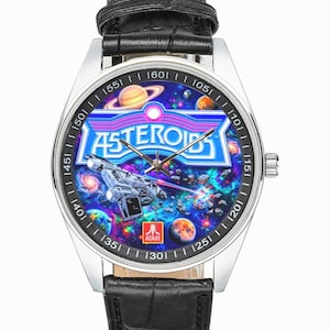 May include: A silver-toned wristwatch with a black leather band. The watch face features a colorful space scene with the word "ASTEROIDS" in a blue and purple font. The Atari logo is visible.