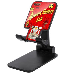 May include: Black adjustable phone stand with a red and yellow graphic that reads "Atomic Energy Lab" with the name "Gilbert" at the top. The stand has a rectangular base and a cradle to hold a phone.