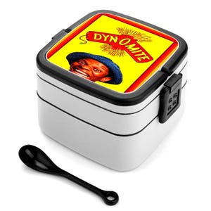 Pode incluir: Lancheira branca de dois níveis com borda e fecho pretos. A tampa apresenta a palavra "DYN-O-MITE" em fundo amarelo, com um homem sorrindo usando um chapéu azul. Uma colher preta está ao lado da lancheira.