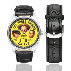 May include: A wristwatch featuring a yellow face with the text "THE THREE STOOGES" and images of Moe, Larry, and Curley. The watch has a silver case and a black leather band. The text "ON TV!" is also visible.